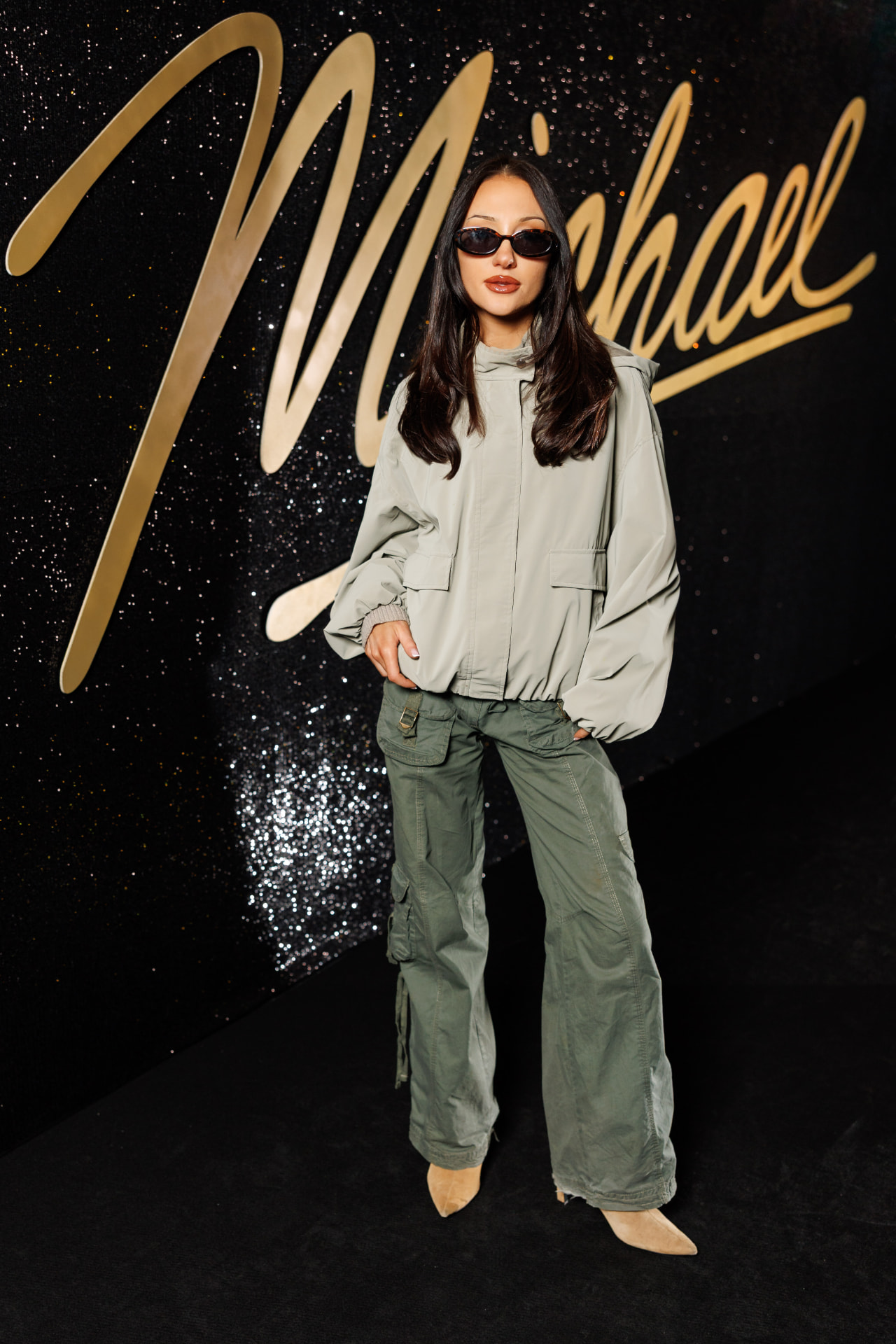 Guest wearing sunglasses, a white jacket and grey trousers in front of a black and gold Michael backdrop