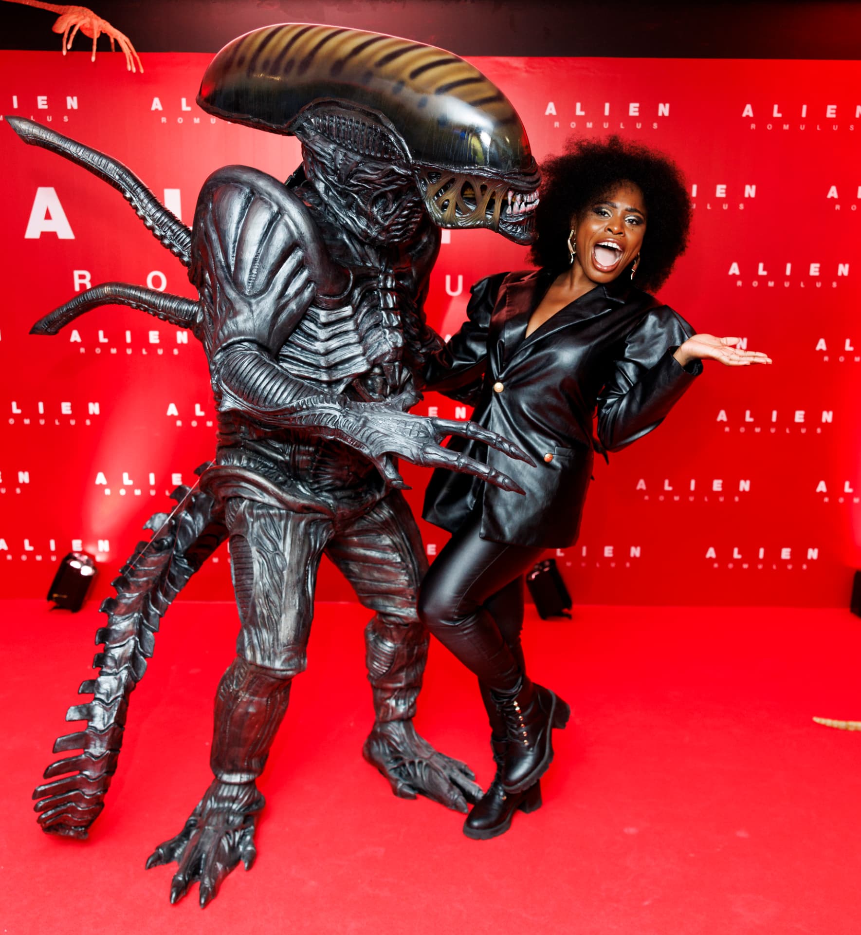 Guest in a black outfit playfully posing beside a costumed Xenomorph against a red Alien: Romulus backdrop