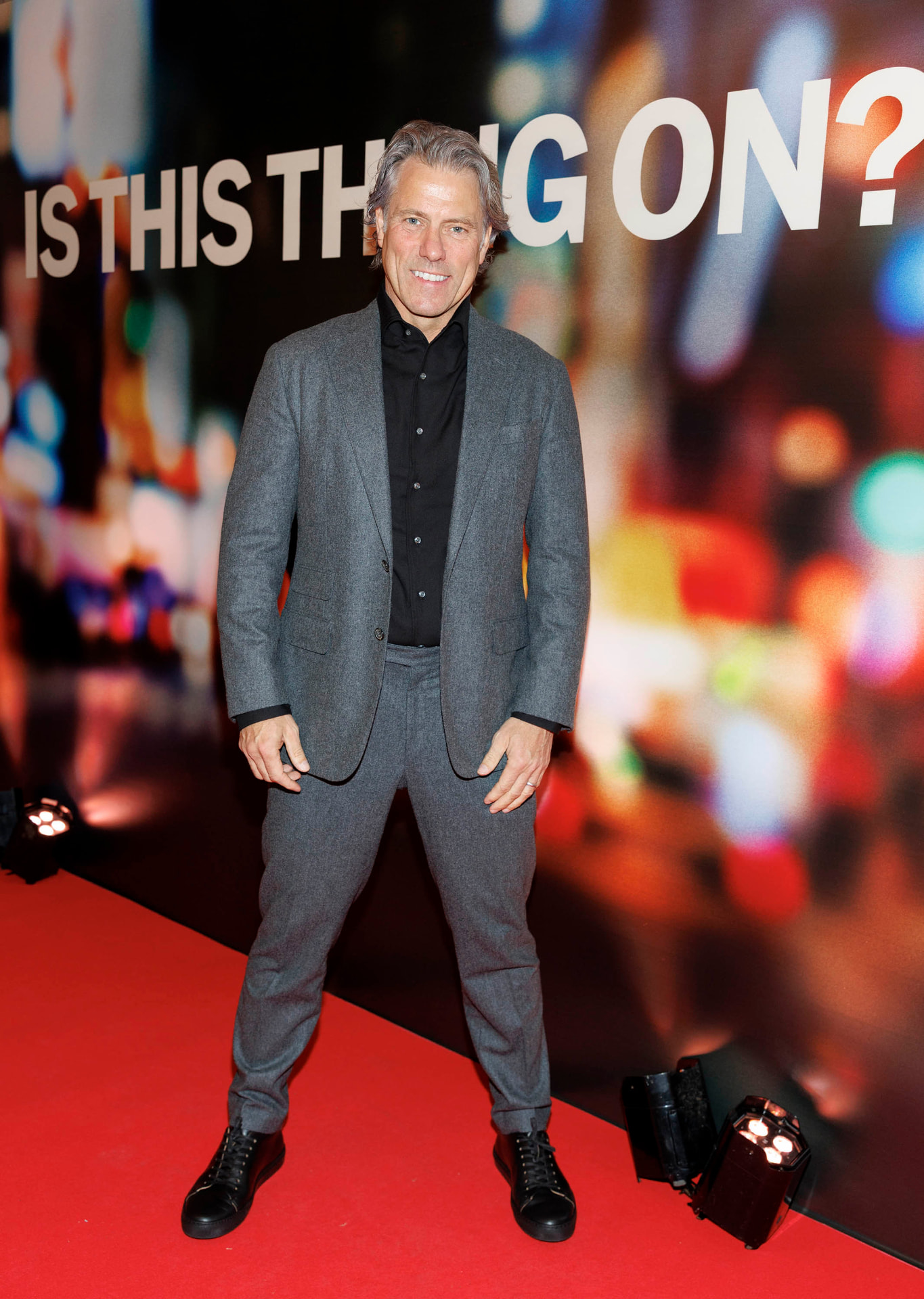 John Bishop smiling in a grey suit on the red carpet in front of an Is This Thing On? backdrop