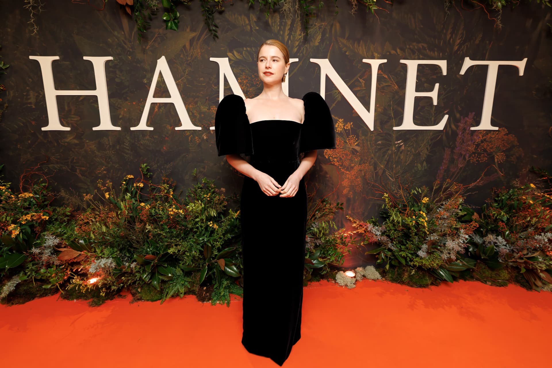 Jessie Buckley in a black off-the-shoulder gown on the red carpet in front of a floral HAMNET backdrop