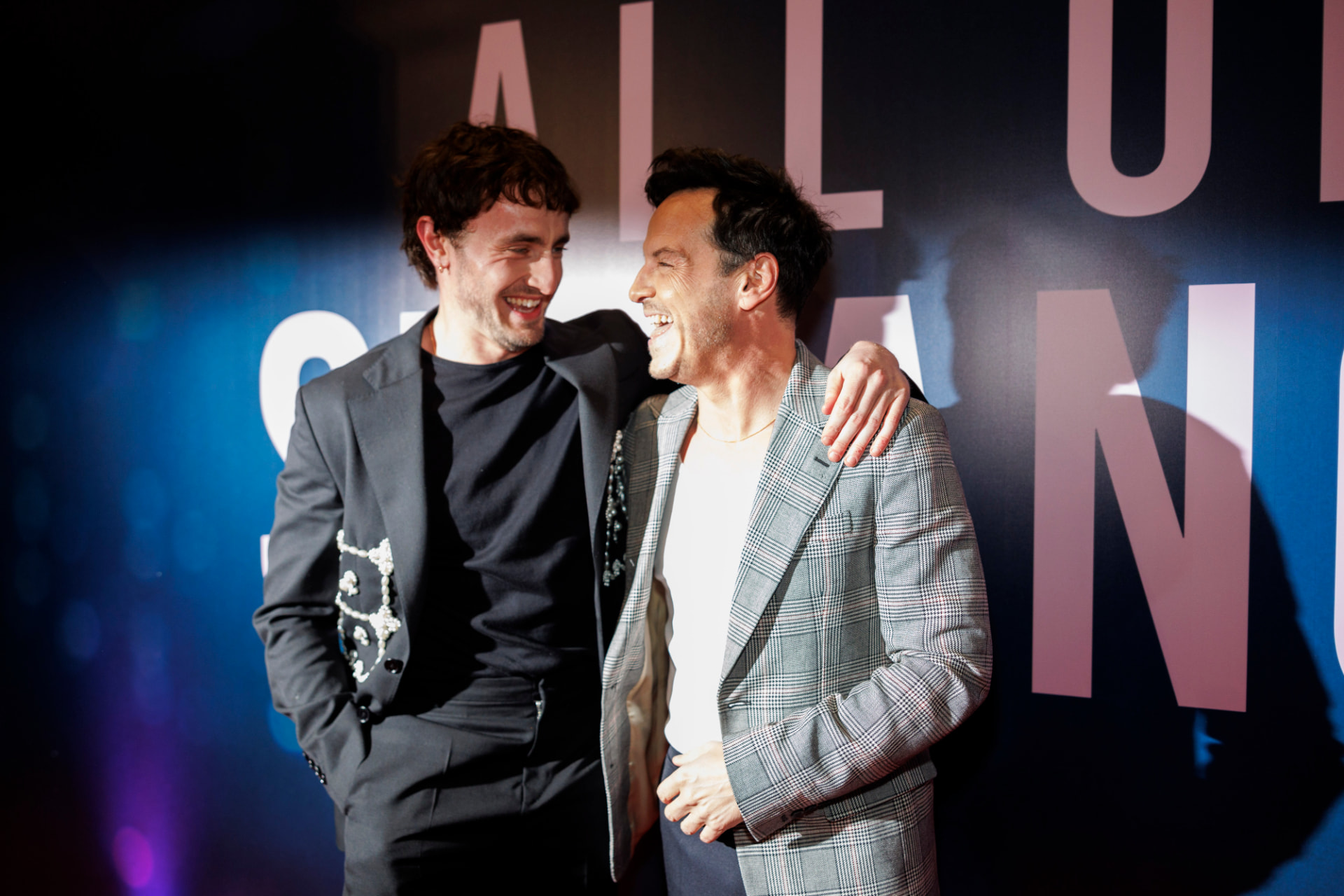 Two actors laughing with an arm around each other in front of the All of Us Strangers backdrop