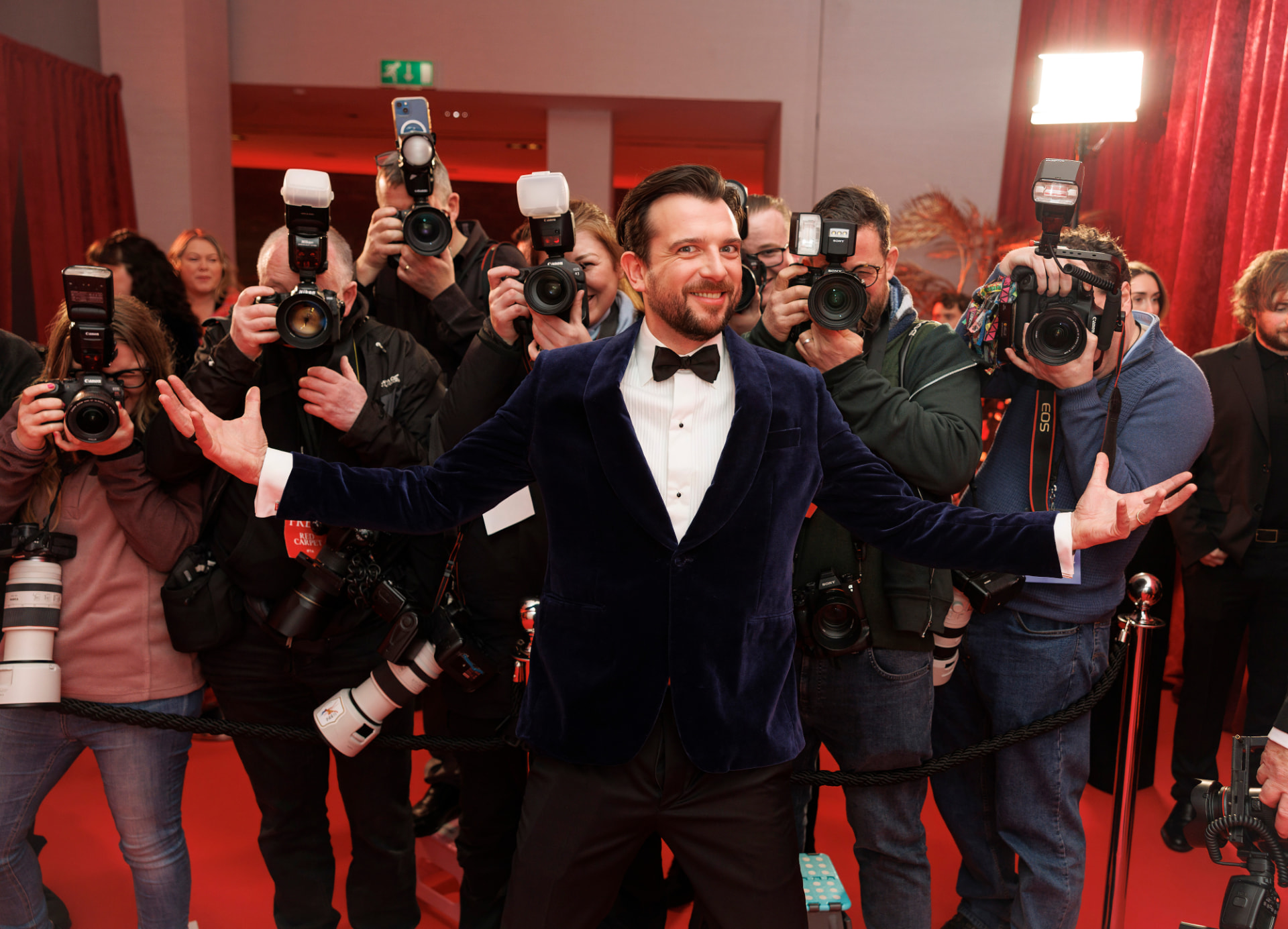 Smiling actor in a blue velvet tuxedo with arms outstretched, surrounded by red-carpet photographers